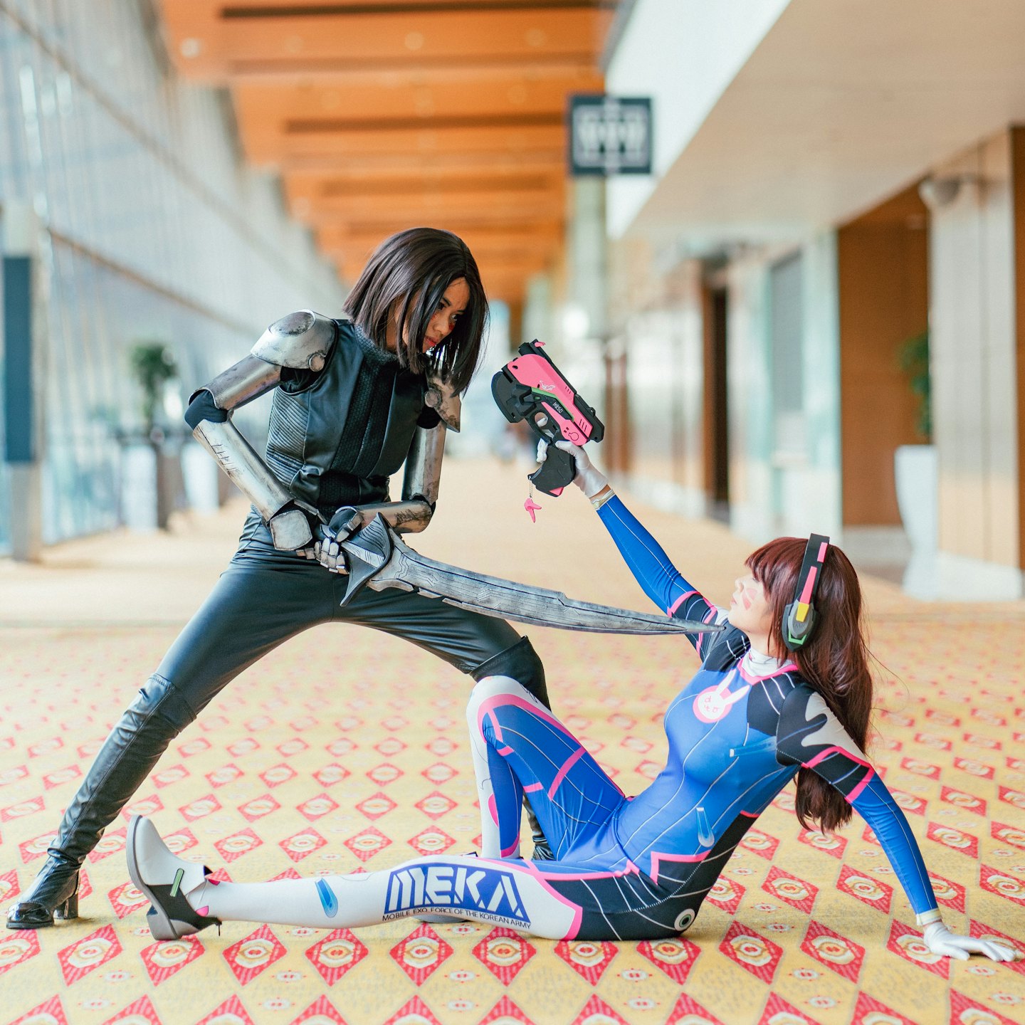 Cosplayers in Alita and D.Va costumes battle with prop weapons.