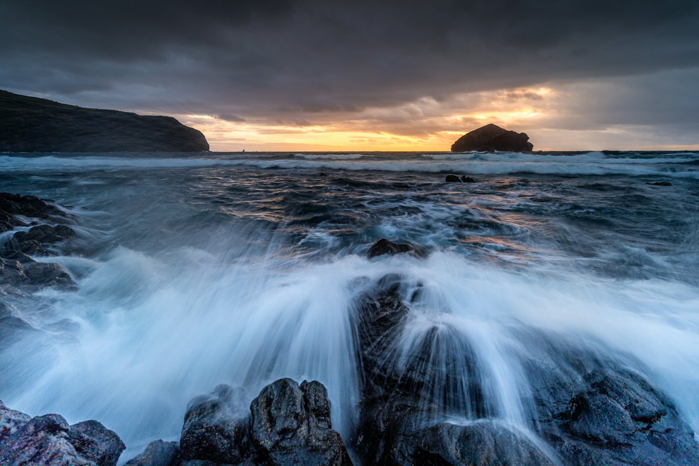 Ocean waves crash on rocky shore at sunset.